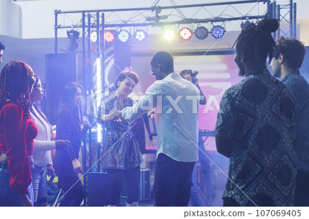 Carefree man and woman couple dancing together with friends at nightclub disco party. Smiling diverse clubbers improvising moves on dancefloor while having fun at club discotheque 107069405