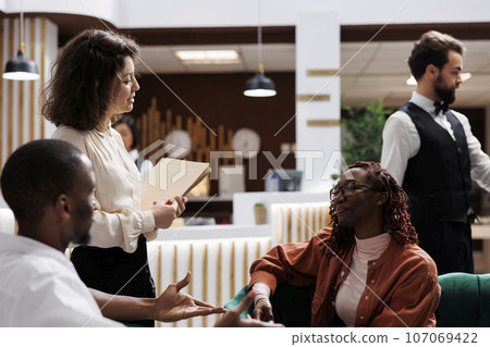 Hotel manager provide service to guests, acting professional in lounge area lobby at luxury resort. Woman working as lodging administrator talking to tourists, reception front desk. Hotel manager provide service to guests, acting professional in lounge area lobby at luxury resort. Woman working as lodging administrator talking to tourists, reception front desk. 107069422