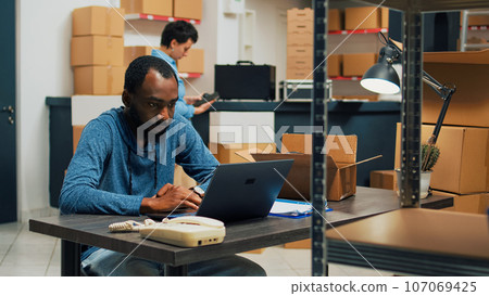 Male entrepreneur planning merchandise inventory and quality control, working on laptop to do retail logistics. Young man using stock in packages, shipping products and doing financial planning. Male entrepreneur planning merchandise inventory and quality control, working on laptop to do retail logistics. Young man using stock in packages, shipping products and doing financial planning. 107069425