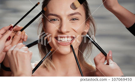 Flawless woman receiving makeover from cosmetologists, team of make up artists and specialist doing natural make up look with brushes. People preparing beauty model for ad campaign. Close up. 107069450