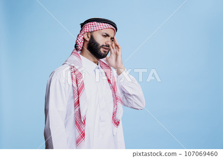 Muslim man wearing traditional clothes suffering from migraine and rubbing temple. Arab person in thobe and headscarf touching head while having headache and posing in studio 107069460
