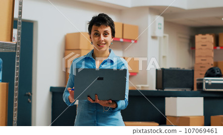 Small business owner using laptop to do storehouse inventory, planning quality control for products before shipping them. Person taking notes on pc and looking at merchandise packaging on shelves. 107069461