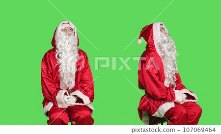 Man dressed as santa claus on chair sitting against greenscreen background, father christmas embodiment. Young adult advertising december seasonal holiday, traditional event. 107069464