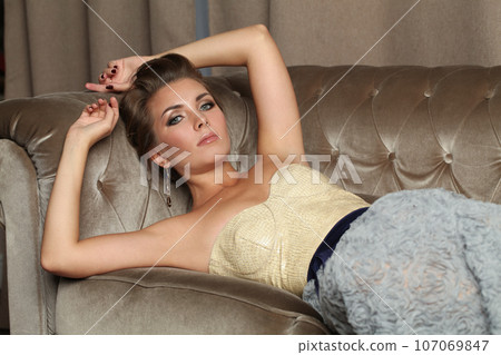Fashion photo of a beautiful young woman in a pretty dress on beige sofa Fashion photo of a beautiful young woman in a pretty dress on beige sofa 107069847