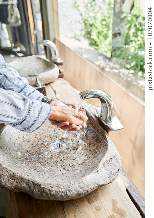 A man's hand touches the water in a beautiful sink with a metal faucet next to an mirror. Close-up of an elegant faucet in the bathroom sink next to stylish decorations. A man's hand touches the water in a beautiful sink with a metal faucet next to an mirror. Close-up of an elegant faucet in the bathroom sink next to stylish decorations. 107070004