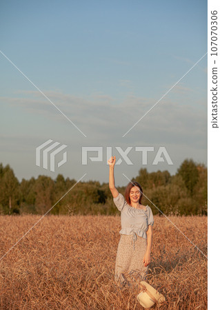 A young beautiful girl with braces on her teeth and long hair poses in a wheat field in the summer at sunset. The girl holds a hat in her hand against the background of a wheat field. 107070306