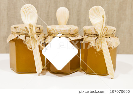 A small yellow jar of honey with a decorative lid made of beige paper and a bow, a small wooden spoon A small yellow jar of honey with a decorative lid made of beige paper and a bow, a small wooden spoon 107070600