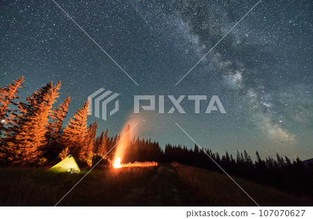 Night camping under sky full of stars and Milky way in the mountains. Starry sky over illuminated tourist tents on hills near forest. Warm light from campfire at dark night. Night camping under sky full of stars and Milky way in the mountains. Starry sky over illuminated tourist tents on hills near forest. Warm light from campfire at dark night. 107070627