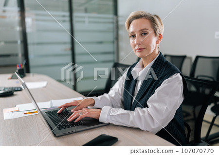 Portrait of elegance middle-aged businesswoman executive top manager sitting at desk working with documents typing on laptop computer, looking at camera with serious expression, in corporate office. Portrait of elegance middle-aged businesswoman executive top manager sitting at desk working with documents typing on laptop computer, looking at camera with serious expression, in corporate office. 107070770