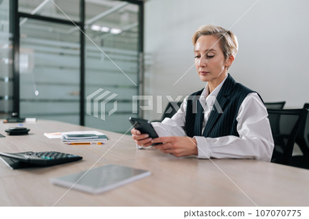 Low-angle view portrait of middle-aged businesswoman using smartphone sitting at office desk touching screen, typing message on mobile phone, browsing Internet, social media, shopping online. Low-angle view portrait of middle-aged businesswoman using smartphone sitting at office desk touching screen, typing message on mobile phone, browsing Internet, social media, shopping online. 107070775