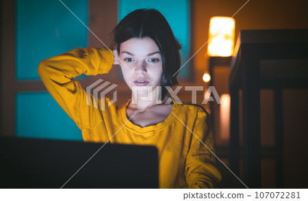 Portrait of beautiful caucasian girl uses laptop while sitting at home. In the evening creative woman works on a computer in her cozy living room. 107072281
