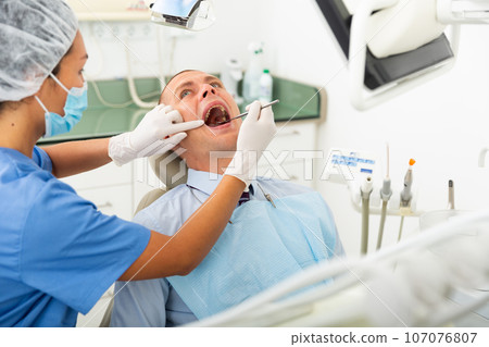 Woman dentist in a protective mask conducts an examines a man patient with the help of tools Woman dentist in a protective mask conducts an examines a man patient with the help of tools 107076807