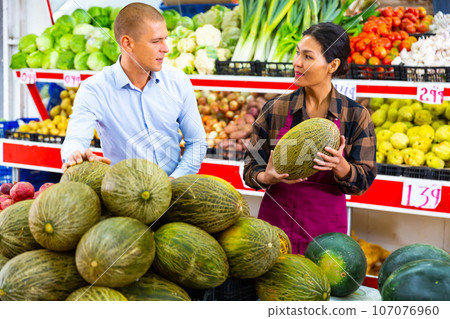 Supermarket worker helping man to choose melon Supermarket worker helping man to choose melon 107076960
