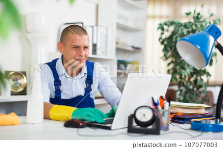 Smiling cleaning service worker sitting at office table with laptop Smiling cleaning service worker sitting at office table with laptop 107077003
