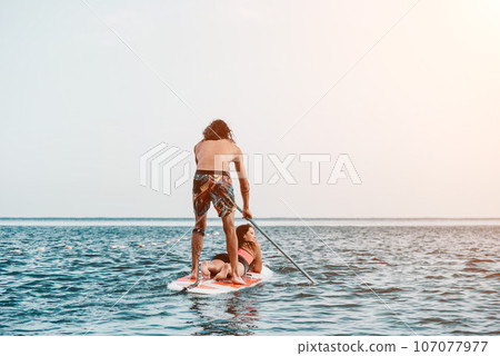 Sea woman and man on sup. Silhouette of happy young woman and man, surfing on SUP board, confident paddling through water surface. Idyllic sunset. Active lifestyle at sea or river. 107077977