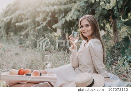 Woman picnic vineyard. Happy woman with a glass of wine at a picnic in the vineyard, wine tasting at sunset and open nature in the summer. Romantic dinner, fruit and wine. 107078110