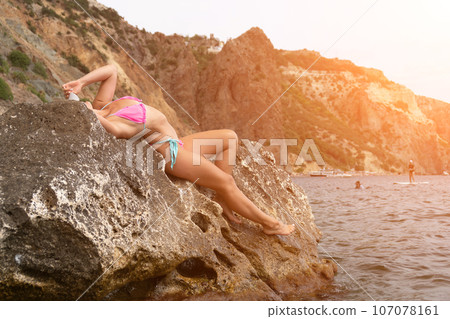 Woman travel sea. Pink bikini tourist captures sea memory, posing on beach amidst volcanic mountains for travel adventure. Happy woman enjoys outdoor photography. Woman travel sea. Pink bikini tourist captures sea memory, posing on beach amidst volcanic mountains for travel adventure. Happy woman enjoys outdoor photography. 107078161