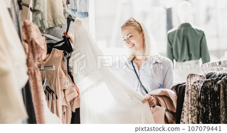 Woman shopping clothes. Female shopper looking at fashionable clothes indoors in clothing store. 107079441
