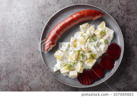 Lightly smoked sausage with a side dish of potatoes with dill and boiled beets close-up on a plate on the table. Horizontal top view 107084881