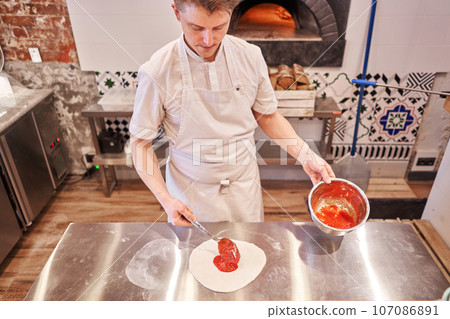 Cooking pizza. the workpiece poured tomato sauce. Closeup hand of chef baker in uniform apron cook at kitchen 107086891