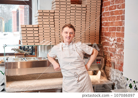 The chef prepares pizza in a neapolitan pizzeria. Cook in a apron in the kitchen with a shovel in his hands. boxes for food delivery on background. 107086897