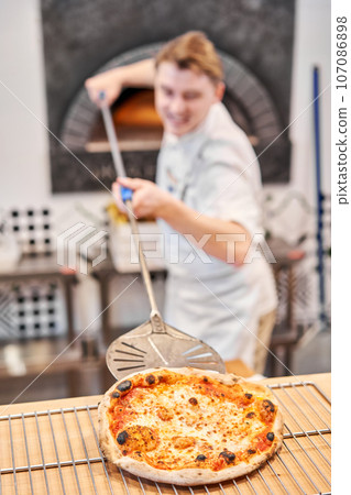 The chef prepares pizza. Raw pizza ready to bake. Cook in a blue apron in the kitchen. with a shovel in his hands. boxes for food delivery on background. The chef prepares pizza. Raw pizza ready to bake. Cook in a blue apron in the kitchen. with a shovel in his hands. boxes for food delivery on background. 107086898