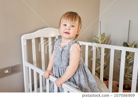 Happy baby girl one and half standing in the crib. Smiling 18 months child playing in sunny bedroom. Baby cot in the parent's bedroom Happy baby girl one and half standing in the crib. Smiling 18 months child playing in sunny bedroom. Baby cot in the parent's bedroom 107086909