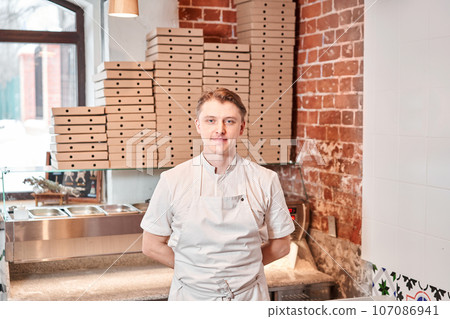 The chef prepares pizza in a neapolitan pizzeria. Cook in a apron in the kitchen with a shovel in his hands. boxes for food delivery on background. 107086941