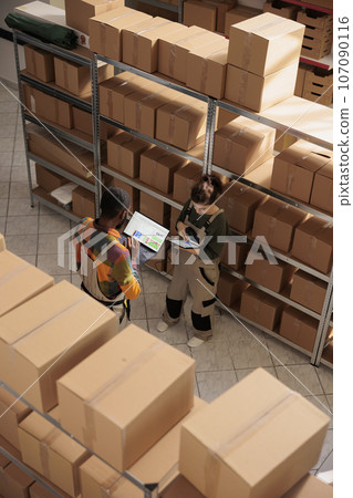 Diverse storehouse team scanning cardboard boxes, checking online products inventory on digital tablet. Storage room employees in protective overalls preparing orders, working in storehouse. Top view 107090116
