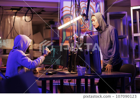 Two hackers discussing cyberattack plan in abandoned warehouse surrounded by technology and hacking equipment. Diverse criminals talking about phishing attack in hideout workspace 107090123