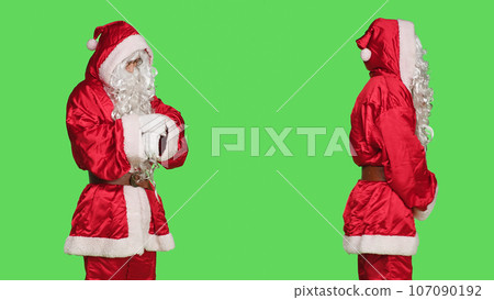 Adult in costume looks at wristwatch to check time against greenscreen backdrop, wearing santa claus red festive suit. Person acting like saint nick to celebrate winter holiday season. 107090192