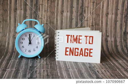 Text, the word Time To Engage is written in a notebook lying on a wooden table with an alarm clock. Text, the word Time To Engage is written in a notebook lying on a wooden table with an alarm clock. 107090667