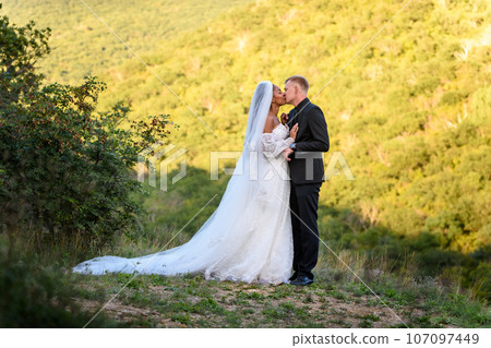 Full-length portrait of the newlyweds against the backdrop of brightly lit foliage, the newlyweds are kissing 107097449
