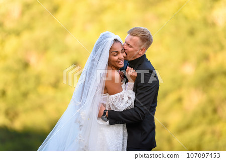Happy newlyweds are hugging against the backdrop of sunny evening foliage, the guy is trying to cheerfully bite the girl 107097453