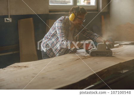 Bearded male wood worker using electric sander for wood. Carpentry workshop. High quality photo 107100548