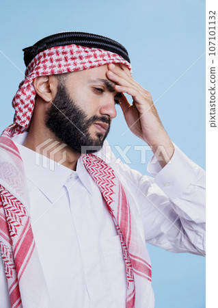 Arab man in ghutra headscarf holding hand on forehead while suffering from headache. Muslim person in pain dressed in islamic thobe and headdress having high blood pressure symptom Arab man in ghutra headscarf holding hand on forehead while suffering from headache. Muslim person in pain dressed in islamic thobe and headdress having high blood pressure symptom 107101132