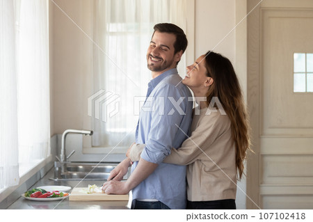 Smiling woman hugging husband from back, standing in modern kitchen 107102418