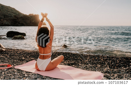 Woman sea yoga. Happy woman in white swimsuit and boho style braclets practicing outdoors on yoga mat by sea on sunset. Women yoga fitness routine. Healthy lifestyle, harmony and meditation 107103617