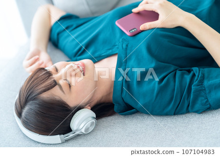 Young woman listening to music on the sofa in the living room while using a smartphone 107104983