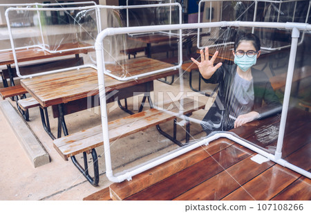 Woman sitting behind a plastic partition (or table shield) setting on table in canteen for reduce and protection the COVID-19 virus spreads in pandemic. Social distancing concept. Woman sitting behind a plastic partition (or table shield) setting on table in canteen for reduce and protection the COVID-19 virus spreads in pandemic. Social distancing concept. 107108266