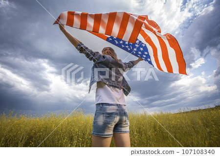 Beautiful young girl holding an American flag on the wind in a rapeseed field. Summer landscape against the blue sky. Beautiful young girl holding an American flag on the wind in a rapeseed field. Summer landscape against the blue sky. 107108340