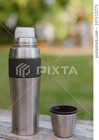 Stainless steel cup, mug with thermos stands on a wooden table in the forest on a green natural background. Thermos and mug with hot drink standing on wet wooden table after rain outdoors, close-up Stainless steel cup, mug with thermos stands on a wooden table in the forest on a green natural background. Thermos and mug with hot drink standing on wet wooden table after rain outdoors, close-up 107113773