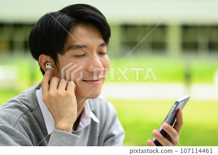Relaxed young asian man listening to music on earphones with closed eyes outside outside campus Relaxed young asian man listening to music on earphones with closed eyes outside outside campus 107114461