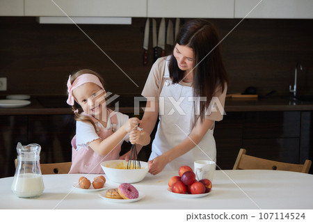 Mother and daughter preparing a sweet cake using flour, milk, sitting on chairs at a table in a modern kitchen. Girl holding a whisk, stirring eggs in a bowl, preparing pancake dough with her mom. Mother and daughter preparing a sweet cake using flour, milk, sitting on chairs at a table in a modern kitchen. Girl holding a whisk, stirring eggs in a bowl, preparing pancake dough with her mom. 107114524