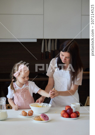Mother and daughter preparing a sweet cake using flour, milk, sitting on chairs at a table in a modern kitchen. Girl holding a whisk, stirring eggs in a bowl, preparing pancake dough with her mom. Mother and daughter preparing a sweet cake using flour, milk, sitting on chairs at a table in a modern kitchen. Girl holding a whisk, stirring eggs in a bowl, preparing pancake dough with her mom. 107114525