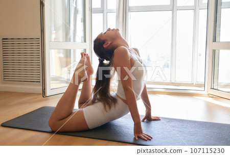Young athletic attractive woman practicing yoga doing exercises. Works out at home or in a yoga studio, sportswear, blue pants and a full-length top indoors. Healthy lifestyle concept. 107115230