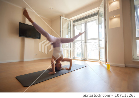 Young athletic attractive woman practicing yoga doing exercises. Works out at home or in a yoga studio, sportswear, blue pants and a full-length top indoors. Healthy lifestyle concept. 107115265