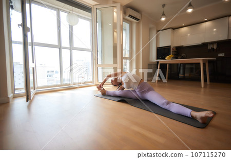 Young athletic attractive woman practicing yoga doing exercises. Works out at home or in a yoga studio, sportswear, blue pants and a full-length top indoors. Healthy lifestyle concept. Young athletic attractive woman practicing yoga doing exercises. Works out at home or in a yoga studio, sportswear, blue pants and a full-length top indoors. Healthy lifestyle concept. 107115270