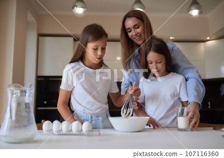 Cute little girl and her beautiful mom in aprons are playing and laughing while kneading the dough in the kitchen. Cute little girl and her beautiful mom in aprons are playing and laughing while kneading the dough in the kitchen. 107116360