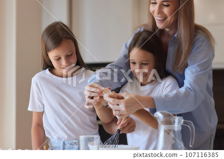 Cute little girl and her beautiful mom in aprons are playing and laughing while kneading the dough in the kitchen. 107116385